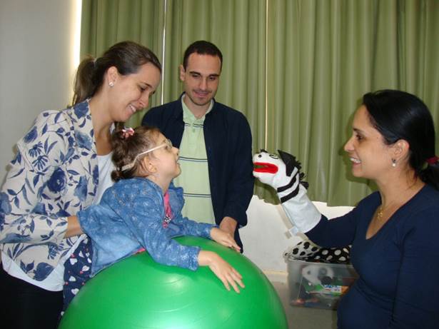 Imagem da terapeuta ocupacional posicionando uma criança com baixa visão sobre uma bola suiça e a sua frente a mãe apresentando fantoche e ao fundo o pai observando. 