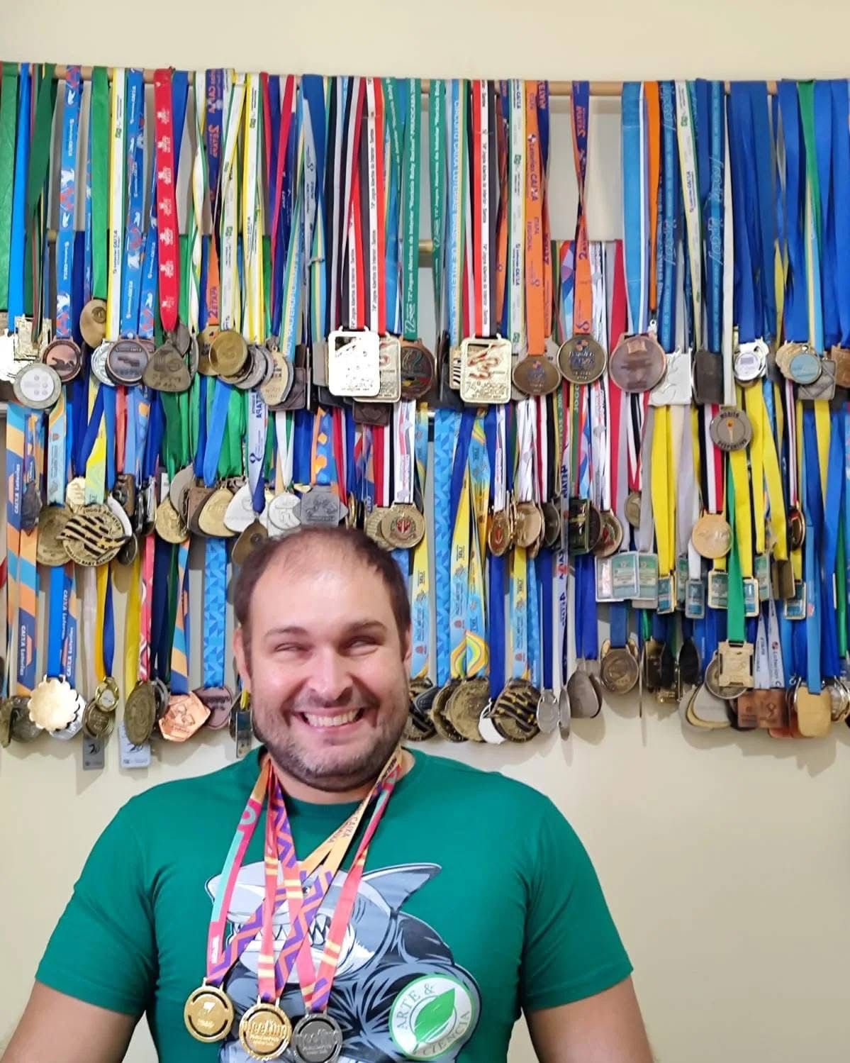 Alex, participante dos programas da PARA-D.V. desde  os 8 anos de idade.  Hoje,  nadador da FUNDESPORT e professor de Educação Física. Na imagem Alex está em pé vestindo uma camiseta verde e ao fundo o seu quadro de medalhas e troféus conquistados em competições municipais, estaduais, nacionais e internacionais. 