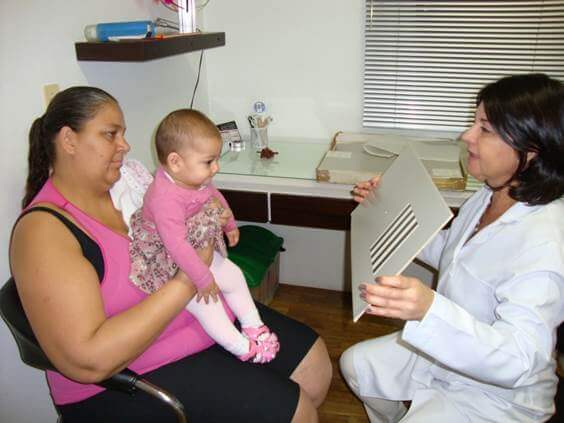 Imagem de um bebê com baixa visão no colo de sua mãe e a sua frente a ortoptista com uma tabela para avaliação da sua acuidade visual de pré-verbais.